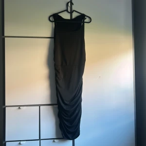 Svart långklänning med drapering - Snygg svart långklänning med smala axelband och draperad nederdel som ger en cool och elegant siluett. Klänningen är ärmlös och har en figurnära passform som framhäver formerna. Perfekt för dig som gillar stilrena och minimalistiska looks.