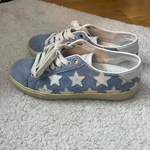 Saint Laurent blå stjärnmönstrade sneakers - Snygga sneakers från Saint Laurent i ljusblå denim med vita stjärnor och vita skinn-detaljer. Skorna har vita snören, rund tå och platt sula. Perfekta för dig som vill sticka ut med en cool och lekfull look.