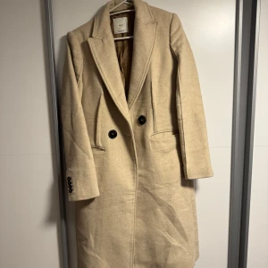 Beige kappa från MNG Suit, XS - Stilren beige kappa från MNG Suit i klassisk modell med två knappar framtill och snygga fickor. Jackan har markerad krage och är helfodrad, perfekt för dig som gillar en clean och tidlös look. Passar dig som vill ha en elegant vibe till höst och vinter.