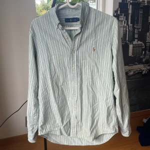 Randig grön skjorta från Ralph Lauren - Snygg grön- och vitrandig skjorta från Ralph Lauren med klassisk krage och broderad logga på bröstet. Skjortan har lång ärm, knäppning framtill och är tillverkad i luftig bomull. Perfekt för en clean och stilren look. 