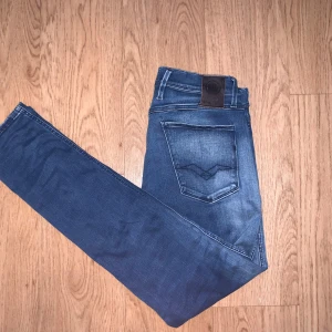 Replay Anbass jeans blå, stl 32/32 - Säljer ett par Replay Anbass jeans i klassisk blå tvätt med snygga slitningar och smal passform. Jeansen har fem fickor, dragkedjegylf och tydlig Replay-logga vid fickan. Perfekta för dig som gillar stilrena och moderna jeans.