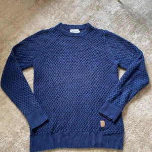 Blå stickad tröja från NN07 - Säljer en blå stickad tröja från NN07 med snyggt strukturerat mönster. Tröjan har rund halsringning, ribbade muddar och lång ärm. Perfekt för dig som gillar stilrena och bekväma plagg med lite extra textur.