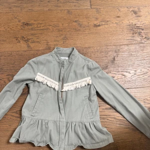 Grön jacka med fransar från Reserved - Snygg ljusgrön jacka från Reserved i bomull med långa ärmar, dragkedja och dekorativa fransar framtill och baktill. Jackan har en peplum-liknande volang nertill och ståkrage för en cool och unik look.