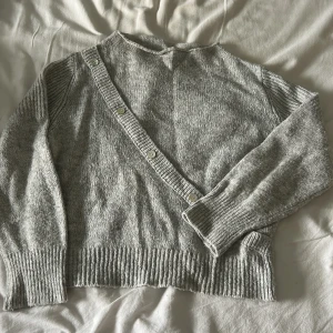 Grå stickad kofta från Design by Si - Mysig grå stickad kofta från Design by Si med snygg omlott-look och knappar i sned rad framtill. Tröjan har ribbade muddar och lång ärm. Materialmix med ull, viskos och syntet för både värme och mjuk känsla. Tröjan är använd men i väldigt bra skick