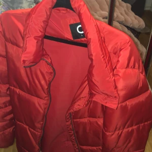 Röd pufferjacka från C - Säljer en röd pufferjacka från C i storlek L. Jackan har en glansig finish, breda vadderade paneler och dragkedja framtill. Perfekt för kalla dagar och ger en riktigt snygg streetstil. Jackan är långärmad och har en hög krage.