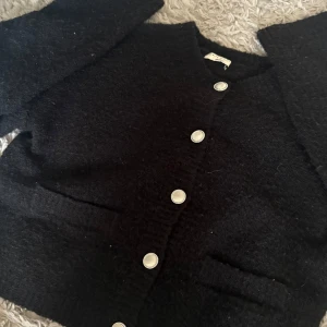 Svart kofta från Gina Tricot - Mysig svart kofta från Gina Tricot med snygga silverfärgade knappar framtill. Koftan har lång ärm, ribbade muddar och två fickor. Perfekt att slänga på sig när det är lite kyligt ute. Materialet är stickat och känns riktigt skönt mot huden.