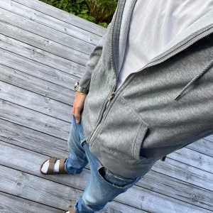 Grå hoodie med dragkedja och huva - Snygg grå hoodie med dragkedja framtill och huva med snörning. Tröjan har två stora fickor och ribbade muddar vid ärmslut och nederkant. Perfekt för chill dagar eller när du vill ha en enkel och stilren look. 200