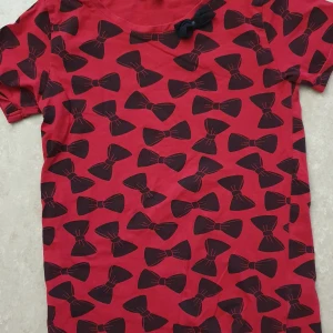 T-shirt - Cool röd t-shirt från mywear young med svart rosettmönster över hela tröjan och en extra svart rosett vid halsen. T-shirten har korta ärmar och är gjord i mjuk bomull, perfekt för dig som gillar unika detaljer och mönster.