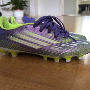 Lila och limegröna adidas fotbollsskor - Snygga fotbollsskor från adidas i lila och limegrönt med vita detaljer. Skorna har snörning, låg profil och mönstrad ovandel i syntetmaterial. Sulan har fasta dobbar i lime och vitt för bra grepp på planen. Perfekta för dig som vill sticka ut på matchen!