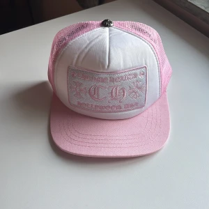 Chrome Hearts rosa och vit keps - Säljer en snygg truckerkeps från Chrome Hearts i rosa och vitt. Kepsen har broderad logga framtill, nät på sidorna och baksidan samt en cool metallknapp på toppen. Perfekt accessoar för att lyfta din stil 👏🤩