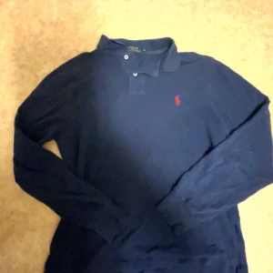 Marinblå långärmad pikétröja Polo Ralph Lauren - Snygg marinblå långärmad pikétröja från Polo Ralph Lauren i slim fit. Klassisk krage, knäppning med två knappar och röd broderad logga på bröstet. Perfekt för dig som gillar stilrena och tidlösa plagg.