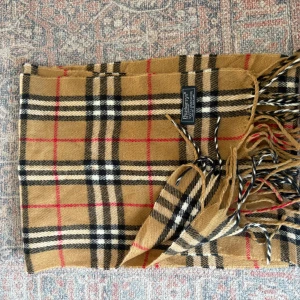Rutig halsduk från Burberry i ull - Säljer en klassisk rutig halsduk från Burberry  i 100% ull. Färgerna är beige, svart, vit och röd med ikoniskt rutmönster och fransar i ändarna. Perfekt accessoar för att lyfta din outfit med lite brittisk vibe.