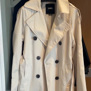Beige trenchcoat från Pull&Bear XS - Stilren beige trenchcoat från Pull&Bear i klassisk modell med dubbelknäppning och stora svarta knappar. Jackan är längre i modellen. Använd 2-3 gånger 