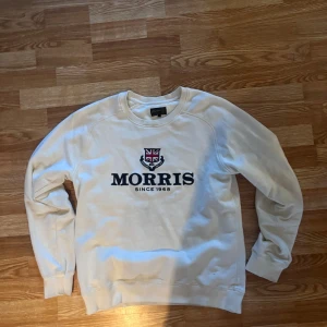 Vit sweatshirt från Morris med tryck - Vit sweatshirt från Morris med stort broderat logotyp-tryck och brittisk flagga på bröstet. Klassisk rund halsringning och långa ärmar. Perfekt för dig som gillar stilren och preppy look.