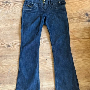 Mörkblå bootcut jeans från River Island - Snygga mörkblå jeans från River Island med bootcut och låg midja. Massa detaljer som dubbla knappar och dragkedjor på bakfickorna. Midjemått 37cm och innerbenslängd 75 cm