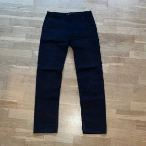 Mörkblå chinos från Lindex, strl 158 - Snygga mörkblå chinos från Lindex i rak modell. Byxorna har klassiska fickor fram och bak, bälteshällor och stängs med knapp och dragkedja. Perfekta för en clean och stilren look.