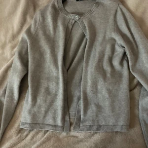 Ljusgrå kofta från Gina Tricot - En stilren ljusgrå kofta från Gina Tricot i storlek L. Koftan har rund halsringning, lång ärm och stängs med en knapp upptill. Tillverkad i mjukt stickat material, perfekt att slänga över en topp för en chill look.