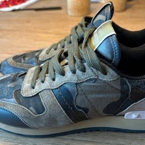 Valentino Rockrunner sneakers camo - Snygga Valentino Rockrunner sneakers med camouflage-mönster i grönt, svart och beige. Skorna har detaljer i mocka och skinn, gröna snören och en röd häl. Perfekt för dig som vill sticka ut med exklusiva och trendiga sneakers.