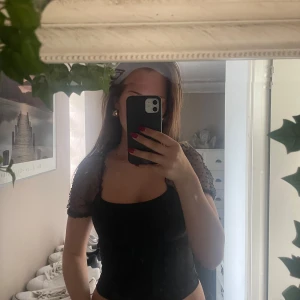 Svart blus - Säljer en svart topp med korta meshärmar som har små prickar. Toppen är figurnära och har en fyrkantig urringning. Ärmarna är genomskinliga och ger en snygg kontrast till den släta kroppen på toppen. Perfekt för dig som gillar trendiga detaljer.