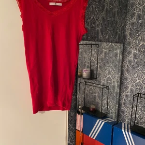 Röd ribbad linnetopp med spets - Snygg röd linnetopp från Gina Tricot med ribbad struktur och spetsdetaljer vid v-ringad hals och axlar. Toppen är ärmlös och har en feminin, figurnära passform som ger en cool vibe. Perfekt att matcha med jeans eller kjol för en trendig look.