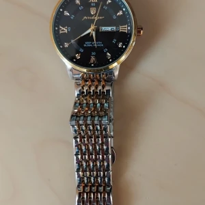 Svart och guld herrklocka Prodragon - Stilren herrklocka från Prodragon med svart urtavla, guldiga romerska siffror och detaljer. Armbandet är i silverfärgad metall med inslag av guld. Klockan har dag- och datumvisning samt gnistrande stenar vid varje timmarkering. Perfekt accessoar för dig som gillar klassisk design.