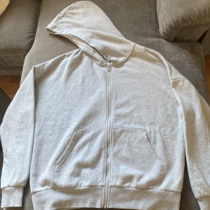 Grå hoodie med dragkedja från Lindex - Enkel och stilren grå hoodie från Lindex med dragkedja framtill och huva. Tröjan har två fickor på magen och är långärmad. Perfekt för chill dagar eller när du vill ha något bekvämt och basic. Materialet är mjukt och skönt, gjord av bomull. Passformen är lite oversized just för mig men kan skilja från person till person