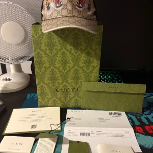 Gucci keps med tigertryck och GG-mönster - Säljer en snygg Gucci keps med klassiskt GG-mönster i beige och svart. Framtill har kepsen två broderade tigrar med öppna käftar och coola detaljer i rött och orange. Baksidan är i svart mesh med justerbart band och Gucci-rand. Perfekt för dig som vill sticka ut.