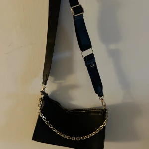 Svart axelväska med guldkedja - Svart axelväska med bred justerbar rem och en guldfärgad kedja som detalj. Väskan har dragkedja upptill och är tillverkad i skinnimitation. 