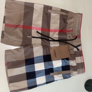 Burberry shorts  - Snygga Burberry shorts med klassiskt rutigt mönster i beige, brun, svart, vit och röd. De har elastisk midja med svart snörning och en bakficka. Materialet är lätt och känns som polyester, perfekt för sommaren. Ikonisk Burberry-tag med London England.