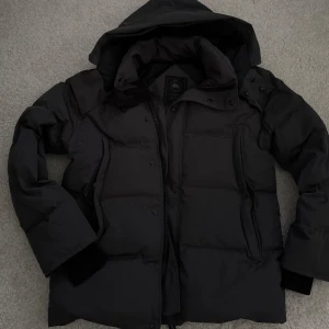 Svart dunjacka från Canada Goose M - Säljer en svart dunjacka från Canada Goose i storlek M. Jackan har huva, dragkedja och knappar framtill samt patch med logga på ärmen. Klassisk pufferstil med quiltade paneler och flera fickor. Perfekt för kalla dagar och riktigt snygg streetstil.