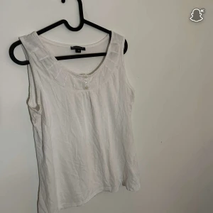 Vit ärmlös linnetopp Brandtex M - Snygg vit linnetopp från Brandtex i storlek M. Toppen är ärmlös med rundad hals och har två dekorativa knappar framtill. Tillverkad i mjuk bomull, perfekt för varma dagar. Enkel och clean design som funkar till jeans eller shorts.