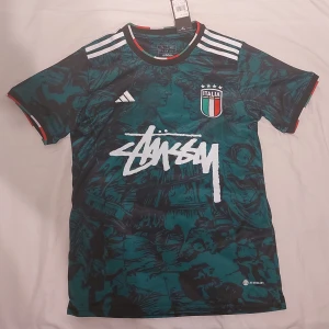 Italien Adidas x Stüssy fotbollströja S/M - Unik Italien fotbollströja från Adidas i samarbete med Stüssy. Tröjan är mörkgrön med svart konstnärligt mönster, vita Adidas-ränder på axlarna och Stüssy-logga i vitt framtill. Ribbade muddar med italienska flaggans färger. Slim fit, officiellt licensierad produkt.