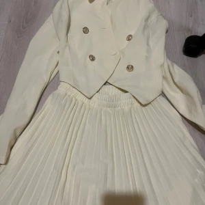 Vit kavaj och plisserad kjol set - Snyggt set med kort vit kavaj med guldknappar och matchande plisserad kjol med resår i midjan. Kavajen har dubbelknäppning och en modern, croppad passform. Kjolen är lång och har fina veck. Perfekt för dig som gillar stilrena och eleganta outfits.