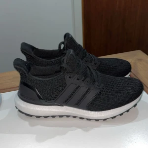 Adidas Ultraboost svarta sneakers - Svarta Adidas Ultraboost sneakers med stickad ovandel och klassiska tre ränder på sidan. Skorna har en vit, dämpad sula och svart yttersula med grovt mönster. Perfekta för dig som gillar sportig stil och vill ha bekväma skor med snörning.