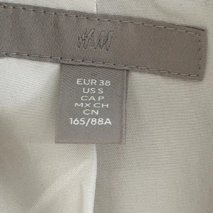 Beige kavaj från H&M, storlek 38 - Snygg beige kavaj från H&M i storlek 38. Klassisk modell med breda slag, två fickor med lock och lätt strukturerat tyg. Perfekt för dig som gillar stilrena och neutrala färger. Passformen är normal och kavajen har en tidlös look.