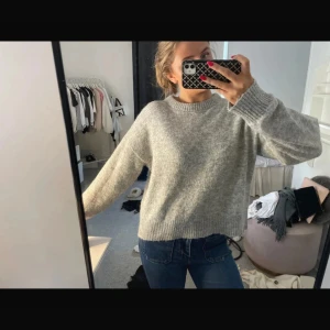 Grå stickad tröja med breda ärmar - Mysig grå stickad tröja med breda, långa ärmar och ribbade muddar. Tröjan har en avslappnad passform och rund halsringning, perfekt att slänga på sig när det är kyligt ute. Enkel och stilren design som funkar till jeans eller kjol.