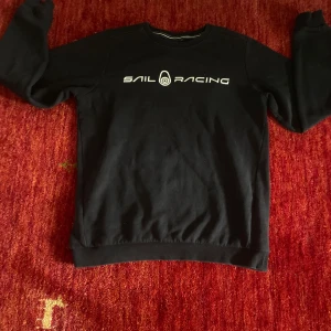 Svart Sail Racing sweatshirt - Svart sweatshirt från Sail Racing med vit logga tryckt över bröstet. Klassisk rund halsringning och långa ärmar. Tröjan är i mjuk bomull och har ribbade muddar vid ärmslut och nederkant. Perfekt för en chill och sportig stil.