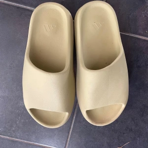 Beige Adidas Yeezy Slides - Säljer ett par beige Adidas Yeezy Slides med minimalistisk design och öppen tå. Tillverkade i syntetmaterial för en mjuk och bekväm känsla. Perfekta för chill dagar eller att glida runt hemma. Det finns ingen box men det är äkta, inte använt på några år.
