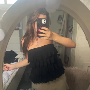 Svart blus - Säljer en svart bandeau tubtopp med flera lager volanger. Toppen är axelbandslös och har en snygg, flowy look som passar perfekt till jeans eller kjol. Materialet känns lätt och luftig, perfekt för varma dagar eller när du vill sticka ut lite extra.