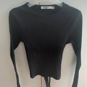 Svart ribbad tröja med öppning i ryggen - Svart långärmad topp från NA-KD med ribbad struktur och öppning i ryggen med  knyts. Toppen har en tight passform och är tillverkad i mjukt material som sitter snyggt. Perfekt för dig som gillar stilrena och trendiga plagg