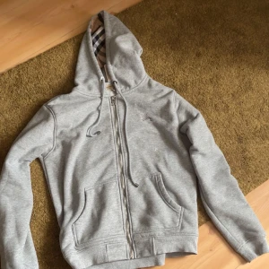 Grå hoodie från Burberry med dragkedja - Grå hoodie från Burberry med klassiskt rutigt foder i huvan. Tröjan har hel dragkedja, snörning i huvan och två stora fickor framtill. Tillverkad i mjuk bomullsmix, perfekt för chill dagar. Ikonisk Burberry-logga broderad på bröstet.