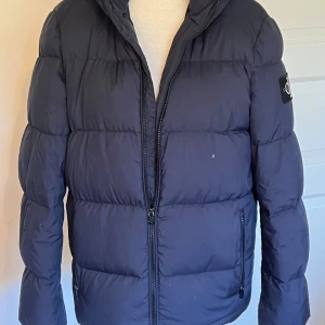 Mörkblå dunjacka från Calvin Klein Jeans - Snygg mörkblå dunjacka från Calvin Klein Jeans med huva och dragkedja framtill. Jackan har puffig design, två fickor med dragkedja och stort CK-logga på ärmen. Perfekt för kalla dagar och riktigt stilren look.