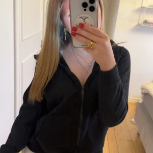 Svart zip-up - En klassisk svart hoodie med dragkedja framtill, huva med snörning och två stora fickor. Tröjan är långärmad och gjord i mjuk bomull, perfekt för chill dagar eller när du vill ha något bekvämt och enkelt.