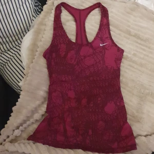Lila-vinröd Nike Dri-Fit linnetopp S - Säljer en lila-vinröd Nike Dri-Fit linnetopp i storlek S. Toppen har ett unikt abstrakt mönster i mörkare röd nyans och klassisk racerback-modell. Perfekt för träning med sitt svala och snabbtorkande material. Liten vit Nike-logga framtill.