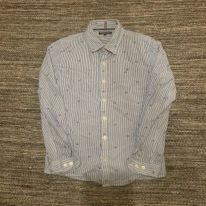 Randig skjorta från Tommy Hilfiger - Snygg blå- och vitrandig skjorta från Tommy Hilfiger med klassisk krage och långa ärmar. Skjortan har broderade loggor och knappar framtill. Tillverkad i mjuk bomull som känns skön mot huden. Passar 150cm lång, om man har funderingar är det bara att skriva!