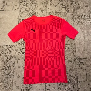 Röd kortärmad fotbollströja Puma M - Snygg röd fotbollströja från Puma i storlek M. Tröjan har ett geometriskt mönster med olika rutor och cirklar på framsidan, samt Puma-loggan i svart på bröstet. Tillverkad i lätt och ventilerande polyester, perfekt för träning eller match.