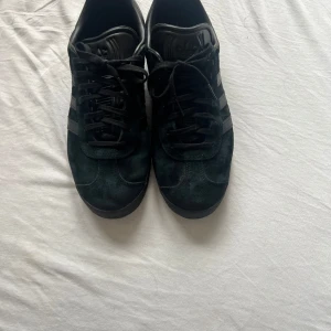 Svarta Adidas Gazelle sneakers mocka - Säljer ett par svarta Adidas Gazelle sneakers i mocka med klassiska tre ränder på sidan och svart sula. Skorna har snörning och rund tå, samt diskret Gazelle-tryck i silver på sidan. Perfekta för dig som gillar stilrena och tidlösa sneakers. Skicket är nästan helt nytt då jag ej använt dem mer än 1-2 gånger. Nypris 1349