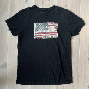Svart t-shirt Ralph Lauren  - Svart t-shirt från Ralph Lauren Denim & Supply i storlek S. Framsidan har ett stort tryck med amerikansk flagga och texten 'Ralph Lauren'. Klassisk rund hals och korta ärmar. 