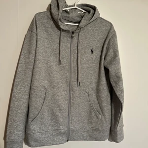 Grå hoodie från Polo Ralph Lauren - Snygg grå hoodie från Polo Ralph Lauren med dragkedja och huva. Tröjan har långa ärmar, två fickor fram och klassisk Polo-logga broderad på bröstet. Mjuk bomullsmix som är perfekt för chill dagar. Den är ALDRIG använd. Jag behöver akut sälja den pga av Sms lån OSV. 