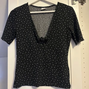 Svart prickig topp från Gina Tricot - Svart topp från Gina Tricot med vita prickar, djup v-ringning och liten rosett framtill. Toppen har korta ärmar och figurnära passform. Perfekt att styla med jeans eller kjol för en trendig look.